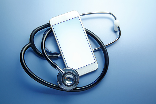A smart phone with a blank screen rests on top of a curled up stethoscope that sits on a blue background.