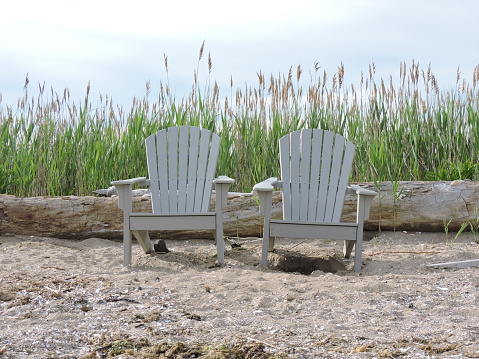 2 Adirondack chairs on empty beach with sand, log  and tall grass.