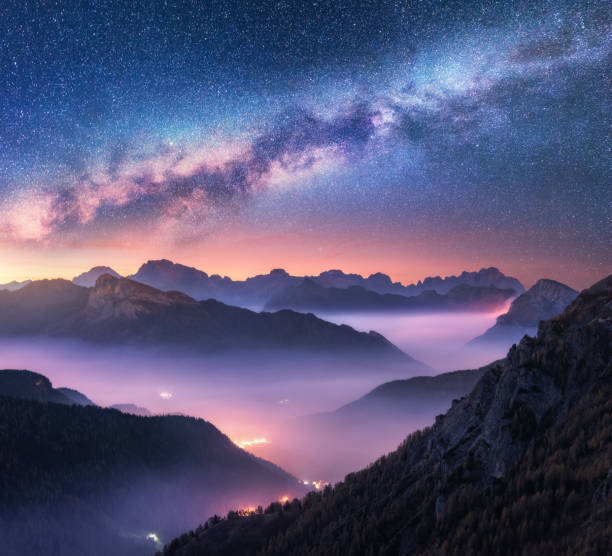 milky way over mountains in fog at night in summer. landscape with alpine mountain valley, purple low clouds, colorful starry sky with milky way, city illumination. passo giau, dolomites, italy. space - nobody aerial view landscape rural scene imagens e fotografias de stock