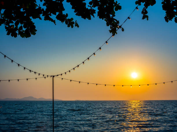 istock Light bulbs hanging on string wire and the sun on sunset sky 1155548730
