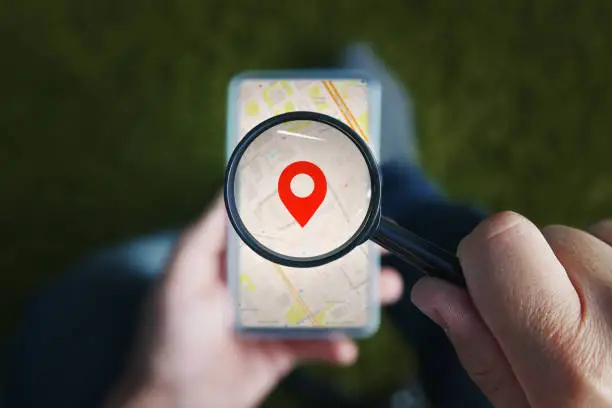 Shot of male hands holding magnifying glass with the red icon of geo-location and looking through it to the smartphone with online city map on the screen. Concept of using mobile net for navigation