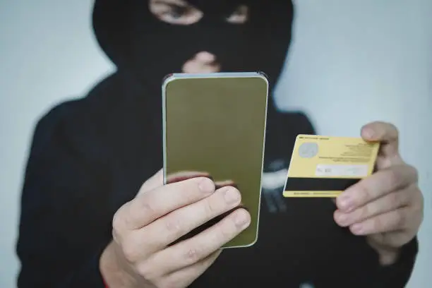 Photo of Cyber criminal in balaclava enters the information of a personal bank account. Credit card fraudulent scheme. Stealing cyber money using mobile. New ways of fraudulent transactions via online banking