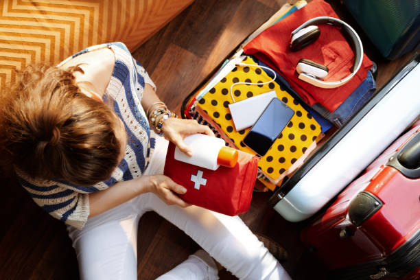 woman packing first aid kit and SPF in open travel suitcase Upper view of modern woman in white pants and striped blouse at modern home in sunny summer day packing first aid kit and SPF in open travel suitcase. first aid kit stock pictures, royalty-free photos & images