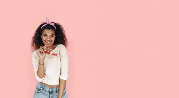 Photo of Happy smiling fashion young african teen girl black woman holding stylish glasses looking at camera isolated on pink summer studio background, banner for website design, portrait, copy space