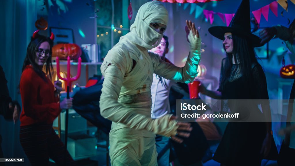 Halloween Costume Party: Old Skinny and Bandaged Mummy Dances. In the Background Zombie, Death, Witch and She Devil Have Fun in a Monster Party Decorated Room Halloween Stock Photo