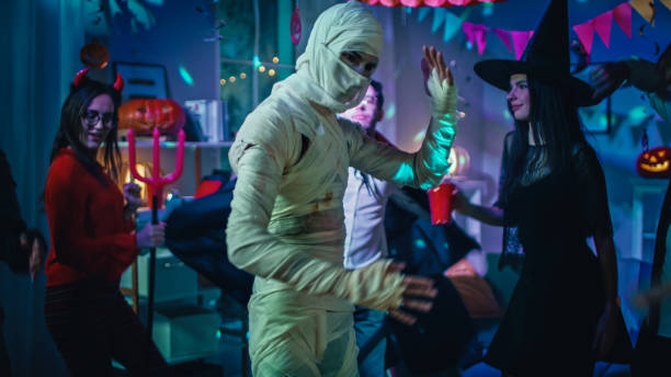 halloween costume party: old skinny and bandaged mummy dances. in the background zombie, death, witch and she devil have fun in a monster party decorated room - halloween horror vampire witch imagens e fotografias de stock