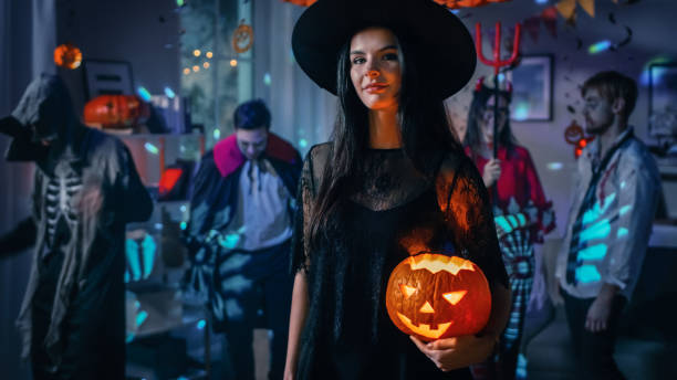 halloween costume party: gorgeous seductive witch wearing dress holds burning pumpkin. background: beautiful devil, scary death, count dracula, zombie dancing in the decorated room - halloween horror vampire witch imagens e fotografias de stock