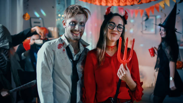 halloween costume party: brain dead zombie i beautiful she devil z trident pose jako para. w tle potwory zabawy i tańca w urządzonym pokoju - costume expressing positivity cheerful close up zdjęcia i obrazy z banku zdjęć
