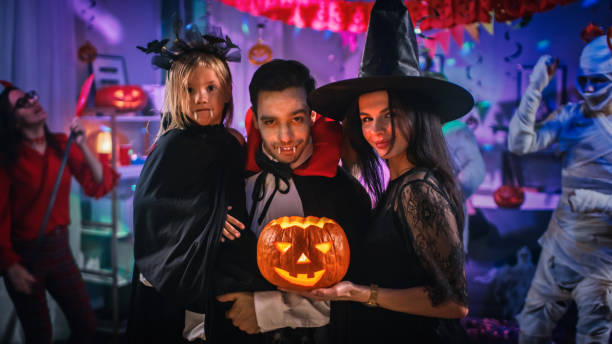 halloween costume party: father count dracula holds little bat girl daughter and hugs witch wife for a happy family portrait. in the background monsters having fun - child party group of people little girls imagens e fotografias de stock