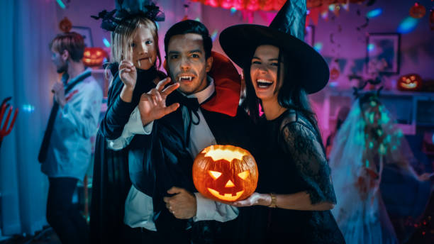 halloween costume party: father count dracula holds little bat girl daughter and hugs witch wife for a happy family portrait. in the background monsters having fun - child party group of people little girls imagens e fotografias de stock