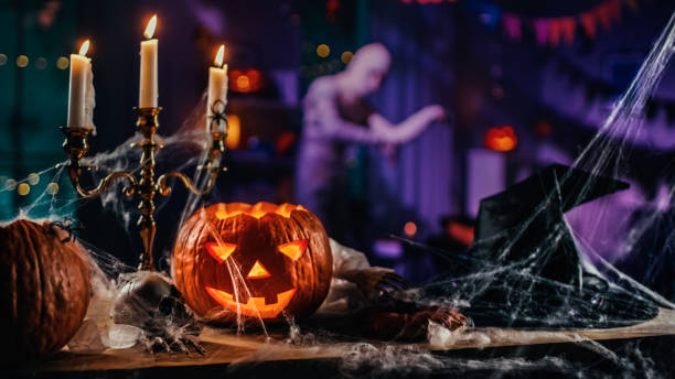 halloween still life colorful theme: scary decorated dark room with table covered in spider webs, burning pumpkin, candlestick, witch's hat and skeleton. in the background silhouette of monster walking by - horror imagens e fotografias de stock