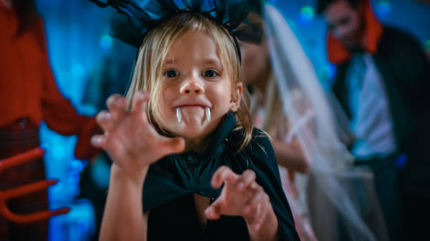 halloween costume party: cute little bat girl with sharp fangs makes scary funny faces. in the background monster party in decorated room and disco lights - child party group of people little girls imagens e fotografias de stock