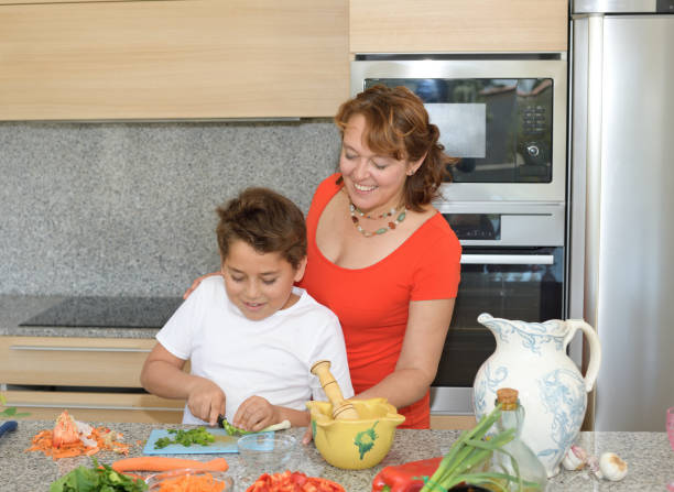 мать и сын готовят обед и улыбаются. ребенок режет чеснок - family with one child domestic life caucasian love стоковые фото и изображения