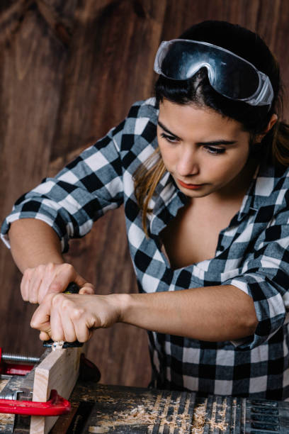 istock Young Hispanic carpenter working with hand plane tool with protective eyewear in home workshop 1155628415