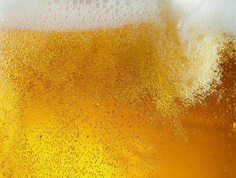 Close up view of floating bubbles in light golden colored beer background. Texture of cooling summer's filtered drink with foam and macro fizz on the glass wall. Fizzing or floating up to top of surface.