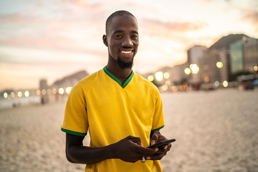 Black man at the beach, looking at camera and holding a phone