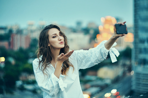 Young beautiful woman making self portrait with her smart phone over cityscape on dusk or video blogging, with evening lightings on background, sending a kiss