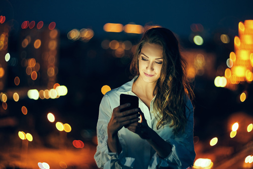 Young woman holding a smart phone in hands texting in the evening with city light on background