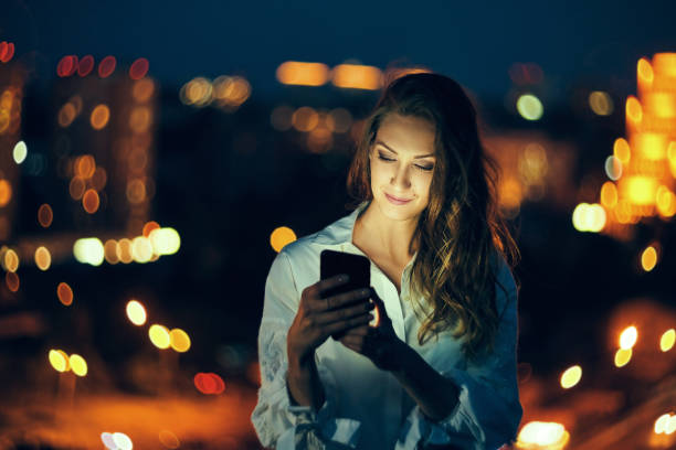 Woman over cityscape holding a smart phone texting Young woman holding a smart phone in hands texting in the evening with city light on background mobile phone lights up stock pictures, royalty-free photos & images