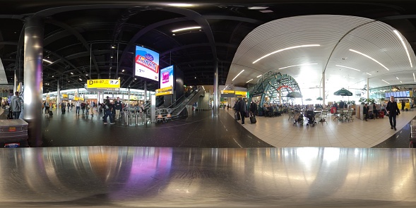 360 degree image taken of the general view inside a departure lounge in the national airport of Schiphol in the city of Amsterdam in the Netherlands. Shows general scene with travellers, shops and restaurants in the walkway area