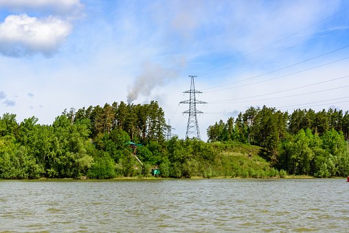 View from the river to the wooded shore and high-voltage tower. Blue sky with clouds on summer day