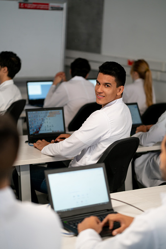 Male student at a computer lab lesson facing camera smiling while working on laptop - Education concepts