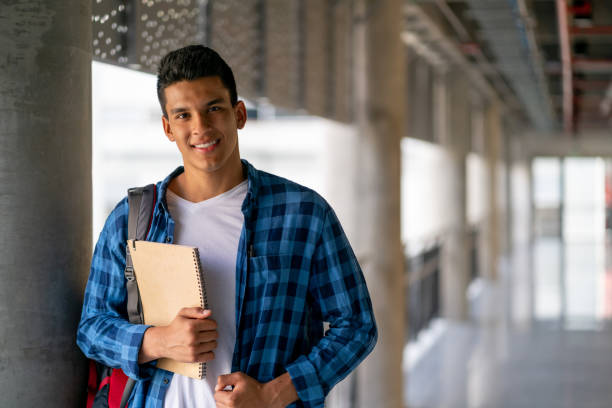 guapo joven estudiante latinoamericano enfrentando cámara sonriendo mientras sostiene cuadernos - men latin american and hispanic ethnicity young men smiling fotografías e imágenes de stock