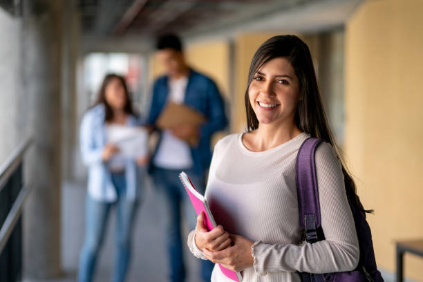 istock Beautiful latin american student facing camera smiling while holding her notebooks and friends standing at background 1155707606