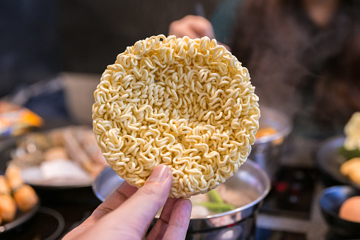 Hand pinching Korean Instant Noodle or dry noodles in circle shape with hot pot table blur background.