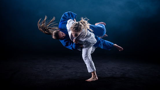 Photo of a woman in a white kimono throwing her sparring partner in a blue kimono to the ground. Black background.