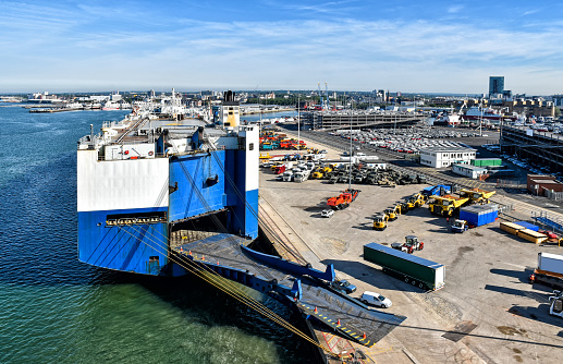 View over the vehicle terminal in the port of Southampton in the United Kingdom. A large vehicle transport ship has moored at the pier. Many vehicles are ready for loading.