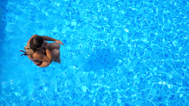 Top view of young woman jumping in pool and splashing crystal clear water. Unrecognizable girl resting and having fun on resort. Lady relaxing during summer vacation or holiday. Slow motion Close up