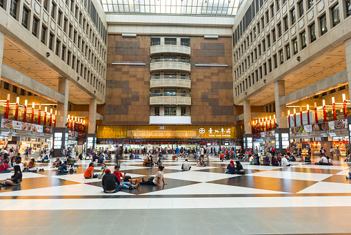 Horizontal color image of the famous ticketing hall of Taipei Main Station in Taiwan. Some people are sitting on the ground while others are walking or on the move. Ticket sales office is visible in the background.