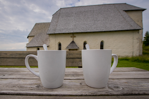 Two white coffe or tea mugs or cups on a wooden table. Church on the top of the hill in the background.