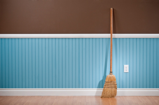 A wooden broom propped up against a wall.  The wall is two different colors.  The top half of the wall is solid chocolate brown.  The bottom half of the wall is bead board, and it has repeating parallel lines spaced a few inches apart.  The bottom section of the wall is a light shade of blue.  Propped up against the wall is a tan wooden broom.  The broom has angled bristles gathered with a dark thread.  There is white molding separating the two sections of the wall and at the floor, which is comprised of light-colored wood.