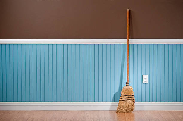 Corn whisk broom standing in empty room A wooden broom propped up against a wall.  The wall is two different colors.  The top half of the wall is solid chocolate brown.  The bottom half of the wall is bead board, and it has repeating parallel lines spaced a few inches apart.  The bottom section of the wall is a light shade of blue.  Propped up against the wall is a tan wooden broom.  The broom has angled bristles gathered with a dark thread.  There is white molding separating the two sections of the wall and at the floor, which is comprised of light-colored wood. sweeping photos stock pictures, royalty-free photos & images