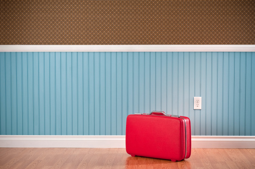 Red vinyl suitcase in empty room. The wall has a blue beadboard wainscoting and a patterned wallpaper.*