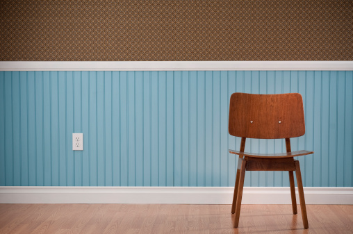 Brown Mid-Century Modern chair in empty room. The wall has a blue beadboard wainscoting and a patterned wallpaper.*