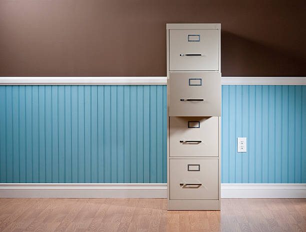 Tan file cabinet, with one drawer open standing in empty domestic room. The wall has a blue beadboard wainscoting, power outlet and a brown painted wall.*