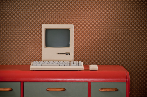 Old classic computer sitting on an art deco retro desk. The wall is covered in a geometric pattern wallpaper.*