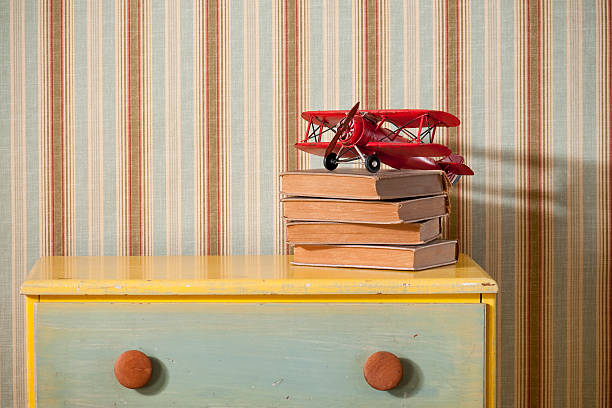 Chest Of Drawers With Books in Empty Bedroom Colorful chest of drawers with old books and toy biplane in empty bedroom. The wall has a striped wallpaper.* dresser stock pictures, royalty-free photos & images