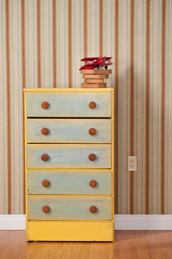 Colorful chest of drawers with old books and toy biplane,  in empty bedroom. The wall has a white baseboard, power outlet  and striped wallpaper.*