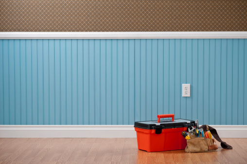 Red toolbox and tool belt in empty domestic room with power outlet. The wall has a blue beadboard wainscoting and a patterned wallpaper.