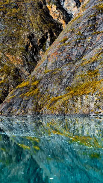 Abstract nature scenery with rocky mountains reflecting in turquoise water and small pieces of ice/icebergs floating in inlet tidewater basin, in Glacier Bay National Park and Preserve, Alaska.