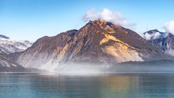 Rocky mountain range in Glacier Bay Alaska with low hanging clouds hovering the peaks and reflection in water. Scenic nature tour sailing through the inlet basin in the National Park and Preserve.