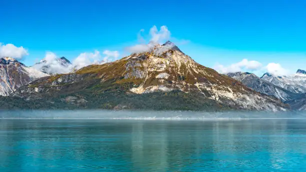 Rocky mountain range in Glacier Bay Alaska with low hanging clouds hovering the peaks and reflection in water. Scenic nature tour sailing through the inlet basin in the National Park and Preserve.