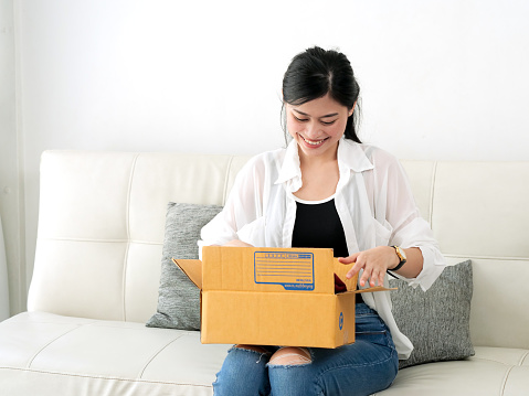 Delivery, mail and people concept - smiling Asian woman opening cardboard box at home.