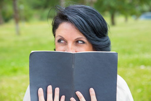 Cheerful sly woman hiding face behind open diary. Middle aged Caucasian lady reading notes outdoors, covering low face with notebook and looking around. Personal diary concept