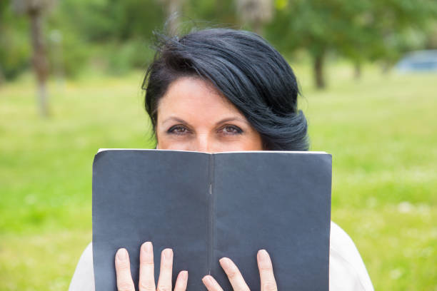 Attractive smiling caucasian blonde female person enjoying reading a story book outdoor. She is sitting in a green meadow field with beautiful sunlight.