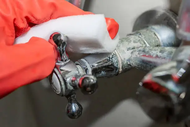 Photo of A red-gloved hand rubs a hot water knob on a chrome-plated faucet covered with limestone with a white melamine sponge. Selective focus. Closeup view